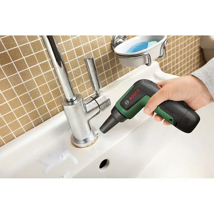 Bosch Akku-Reinigungsbürste Taydo Set - 3,6 V, 1 Akku, 15 -tlg. - Eine Hand hält eine grün-schwarze Bosch TAYDO elektrische Reinigungsbürste, die aktiv um einen Chromhahn in einem weißen Waschbecken mit Schaum schrubbt.