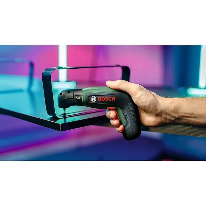 Bosch Akkuschrauber IXO 8 - 4 V, 1 Akku, 2 Ah, 6,5 Nm, 14 -tlg. - Eine Hand hält einen grün-schwarzen Bosch IXO Akkuschrauber, bereit, eine Schraube in eine schwarze Fläche zu drehen, die eine Halterung befestigt.