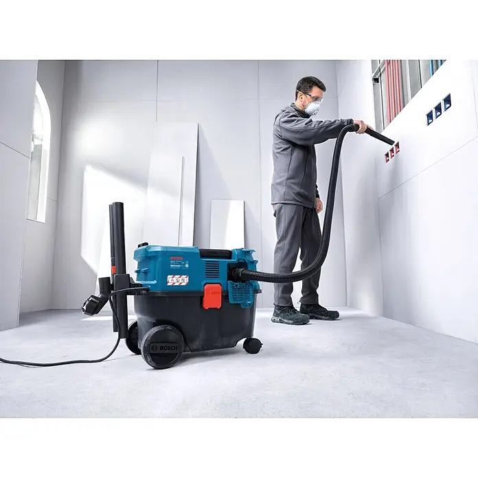 Bosch Professional Elektro-Nass- & Trockensauger GAS12-25LS - 1.200 W, Behältervolumen: 25 l, Halbautomatisch - Ein Arbeiter nutzt einen blau-schwarzen Bosch Sauger, um Schmutz aus Wandausschnitten auf einer Baustelle zu entfernen.