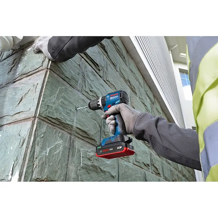 Bosch Professional AMPShare 18V Akku-Schlagbohrschrauber - 18 V, Ohne Akku, 70 Nm, Ohne L-BOXX 136 - Blaue und schwarze Bosch Akkubohrmaschine mit rotem 18V Akku bohrt in strukturierte Schieferwand.