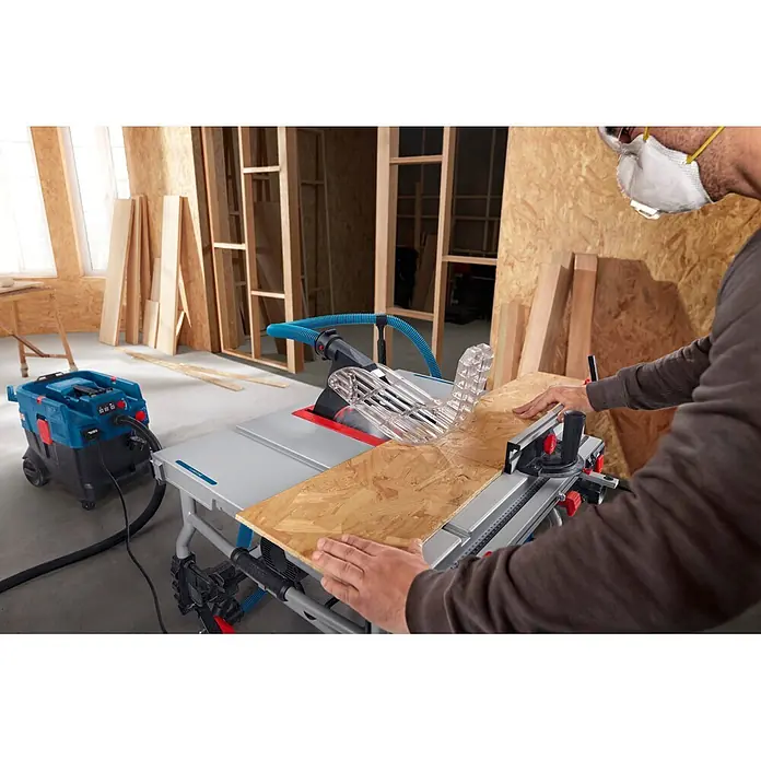 Bosch Professional Tischkreissäge GTS100-254 - 2.200 W, Durchmesser Sägeblatt: 254 mm, Schnitttiefe bei 90 °: 100 mm - Eine Person mit Atemschutzmaske bedient eine blau-silberne Tischkreissäge zum Holzzuschnitt, verbunden mit einem Staubsauger.
