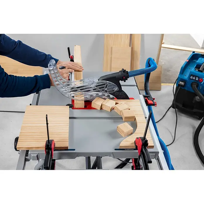 Bosch Professional Tischkreissäge GTS100-254 - 2.200 W, Durchmesser Sägeblatt: 254 mm, Schnitttiefe bei 90 °: 100 mm - Person sägt Holz auf einer Tischkreissäge mit transparentem Schutz und blauem Absaugschlauch.