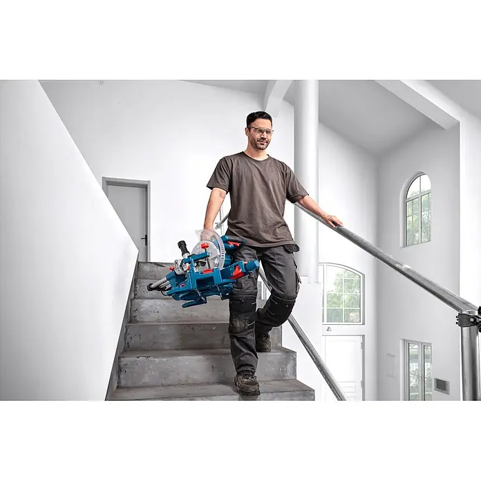 Bosch Professional Kapp- & Gehrungssäge GCM305-216S - 1.300 W, Durchmesser Sägeblatt: 216 mm, Leerlaufdrehzahl: 4.320 U/min - 5.280 U/min - Mann in Arbeitskleidung trägt eine blaue und rote, tragbare Kappsäge auf Treppen.