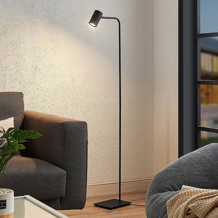 Lindby Stehleuchte Sanad - schwarz, schwarz, GU10 - Eine schlanke schwarze Stehlampe aus Metall mit Spotlichtkopf steht neben einem Sofa und beleuchtet eine strukturierte Wand.