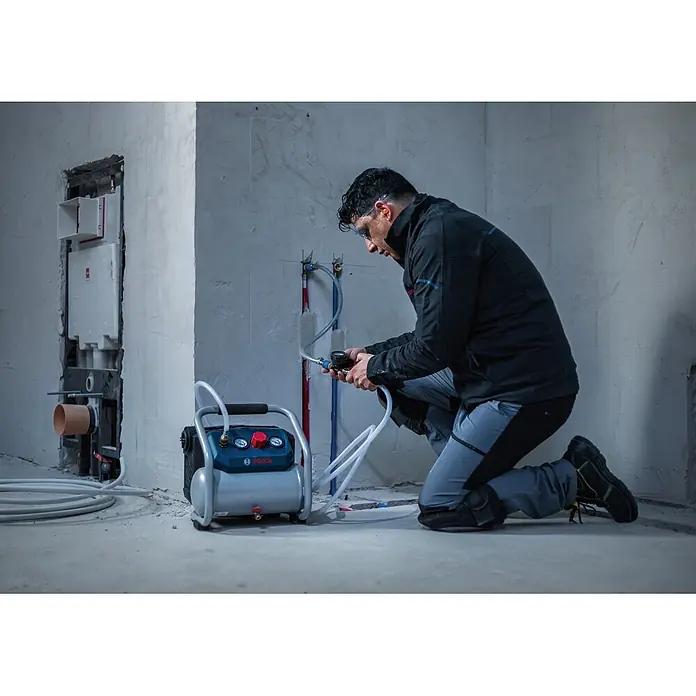 Bosch Professional AMPShare 18V Akku-Kompressor GKO18V-51 - 18 V, 8 bar - Grauer und blauer Bosch Luftkompressor, von einem Arbeiter zum Drucktesten von roten und blauen Sanitärrohren in einem Rohbau verwendet.