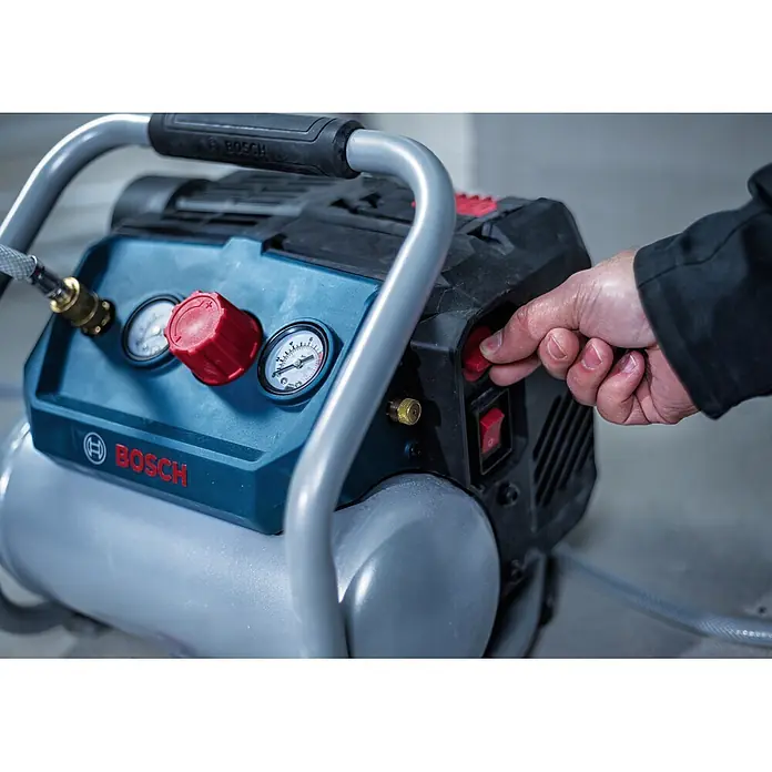 Bosch Professional AMPShare 18V Akku-Kompressor GKO18V-51 - 18 V, 8 bar - Eine Hand betätigt den roten Einschaltknopf eines blau-schwarzen Bosch Kompressors mit sichtbaren Manometern und einem silbernen Tank.