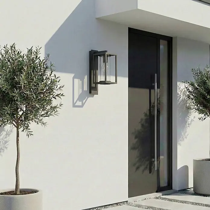Lucande Außenwandleuchte Ferda - schwarz, schwarz, IP44 - Moderne Außenwandleuchte aus dunklem Metall und Glas, die den Eingang eines modernen Hauses beleuchtet, flankiert von Olivenbäumen.
