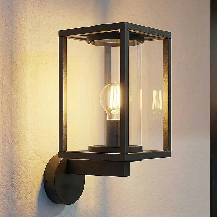 Lucande Außenwandleuchte Ferda - schwarz, schwarz, IP44 - Schwarze rechteckige Wandleuchte mit klarem Glaszylinder und sichtbarer Glühbirne, an einer strukturierten Wand montiert.