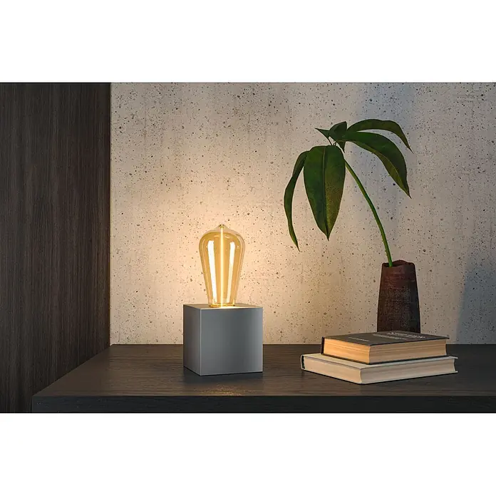 Paulmann LED-Lampe Floating ST64 Gold - E27, Warmweiß, 140 lm, 2,8 W - Dunkelgraue Würfel-Tischlampe mit leuchtender Edison-Glühbirne, die eine dunkle Holzoberfläche neben Büchern und einer Pflanze beleuchtet.