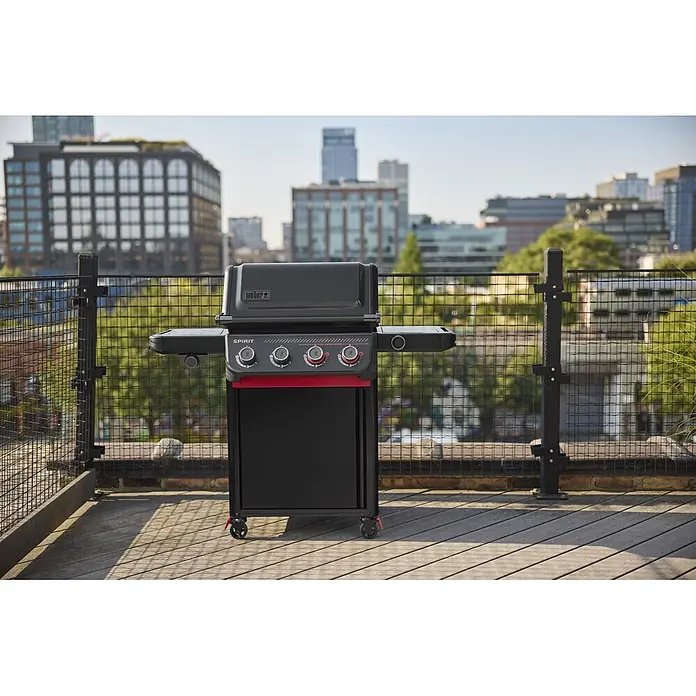 Weber Barbacoa de gas Spirit EPX-435R + Rustidor - Superficie de la parrilla (An x Pr): 61 x 45 cm, 10,3 kW, Número de quemadores: 4 - Parrilla Weber Spirit negra con detalles rojos en una terraza de madera en la azotea, con la ciudad de fondo.