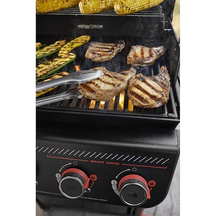 Weber Barbacoa de gas Spirit EPX-435R + Rustidor - Superficie de la parrilla (An x Pr): 61 x 45 cm, 10,3 kW, Número de quemadores: 4 - Parrilla de gas negra sellando chuletas de cerdo, calabacín y mazorcas de maíz, con llamas visibles y pinzas girando una chuleta en la parrilla.