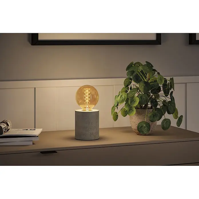 Paulmann LED-Lampe S-Form G95 - E27, Dimmbar, Warmweiß, 300 lm, 4,2 W - Beton-Tischlampe mit glühender Edison-Birne, beleuchtet einen Nachttisch neben einer Pflanze.