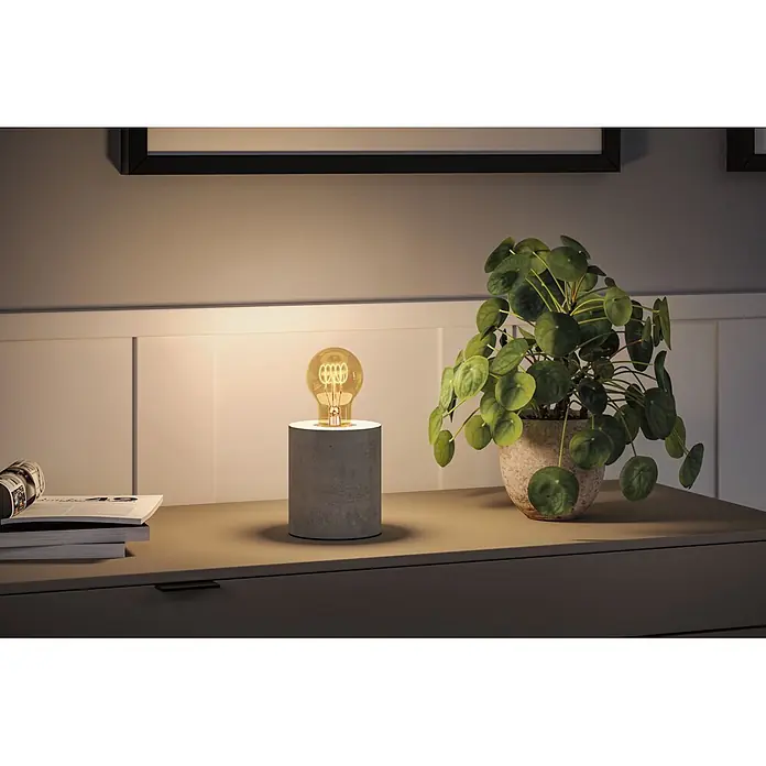 Paulmann LED-Lampe Birne AGL - E27, Dimmbar, Warmweiß, 300 lm, 4,2 W - Eine minimalistische Tischlampe mit grauem zylindrischem Betonsockel und freiliegender Edison-Glühbirne beleuchtet eine Kommode neben einer Pflanze und Büchern.