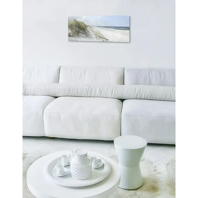 Glasbild - Sommertag III, B x H: 80 x 30 cm - Ein helles Wohnzimmer mit weißem Sofa, Tisch, Hocker und einem gerahmten Bild an der Wand, das ein ruhiges Strandes zeigt.
