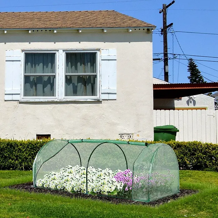 Outsunny Gewächshaus - 100 x 200 cm, Material Seitenwand: Kunststoff, 0,3 mm, Grün - Ein kleines, grünes Netz-Gewächshaus bedeckt weiße und violette Blumen in einem Gartenbeet vor einem Haus.