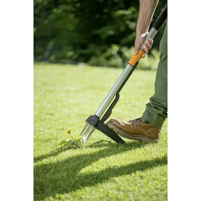 Fiskars Teleskop-Unkrautstecher SmarFit - Länge Stiel: 99 cm - 119 cm - Nahaufnahme eines Fiskars-Unkrautstechers in Aktion, der auf einem Rasen mit einem Stiefel bedient wird.