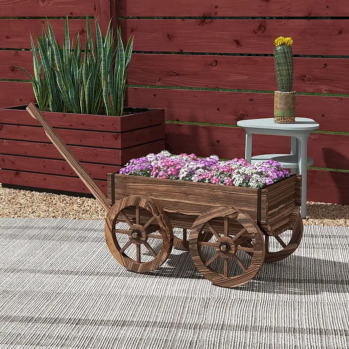 Furnicato Hochbeet - 120 x 41 x 54 cm, Holz, Verkohlt - Hölzerner Wagen-Pflanzkasten gefüllt mit rosa und lila Blumen, neben einem großen Holzpflanzkasten mit Bogenhanf auf einem Terrassenteppich.