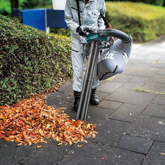 Makita XGT 40V max. Akku-Gebläse UB003GZ - 40 V, Ohne Akku, Blasgeschwindigkeit: 68 m/s - Makita Laubbläser/Sauger in Türkis/Schwarz, von Person auf Weg zum Entfernen von Blättern genutzt. Grauer Auffangsack ist befestigt.