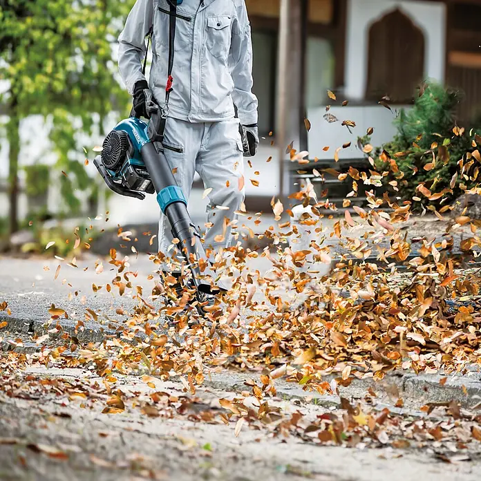 Makita XGT 40V max. Akku-Gebläse UB003GZ - 40 V, Ohne Akku, Blasgeschwindigkeit: 68 m/s - Person bedient einen türkis-schwarzen Laubbläser im Freien, der Herbstlaub auf einem gepflasterten Weg bläst.