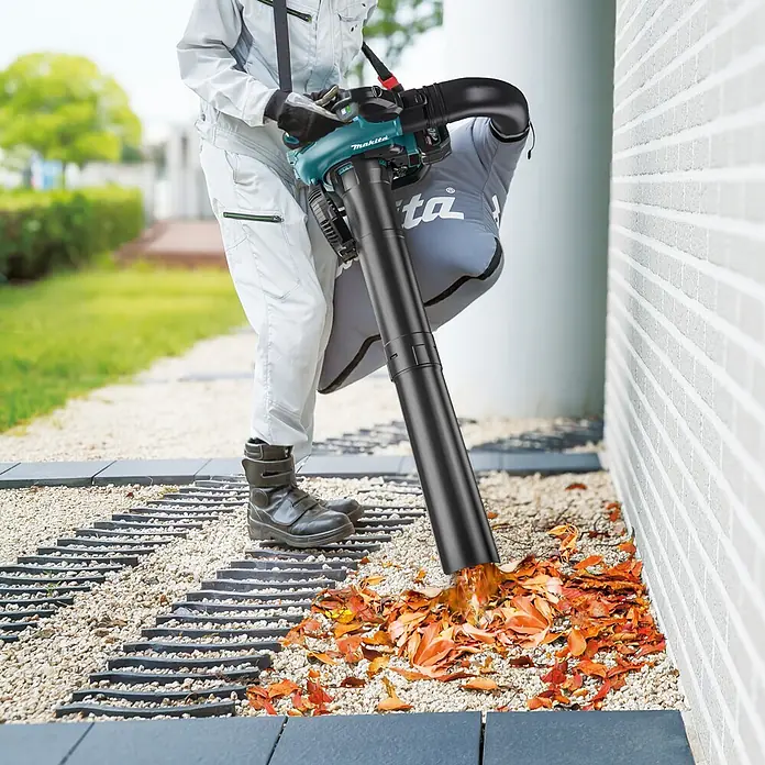 Makita XGT 40V max. Akku-Gebläse UB003GZ - 40 V, Ohne Akku, Blasgeschwindigkeit: 68 m/s - Person in Arbeitskleidung verwendet einen grün-schwarzen Makita Laubbläser/-sauger, um Herbstlaub von einem Kiesweg zu entfernen.
