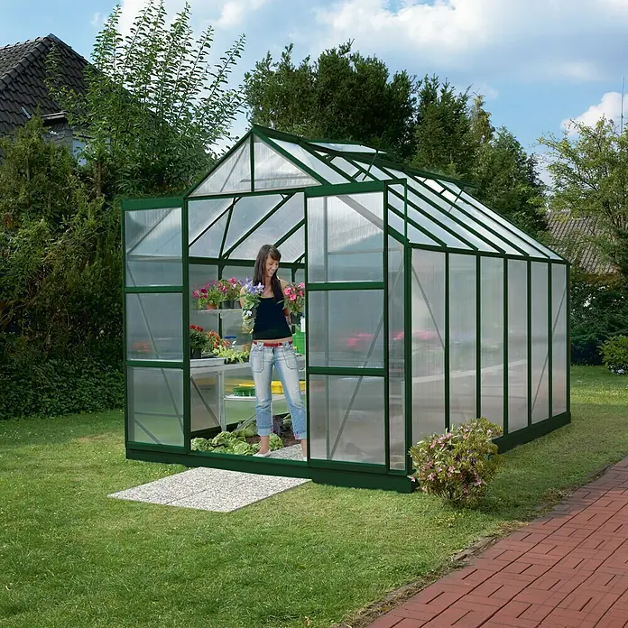Vitavia Gewächshaus Mars 9900 Plus - Außenmaß inkl. Dachüberstand (B x T): 257 x 383 cm, 6 mm, Smaragd - Eine Frau betritt ein grünes Gewächshaus mit durchsichtigen Wänden, in dem Pflanzen auf Regalen stehen, umgeben von einem grünen Garten.