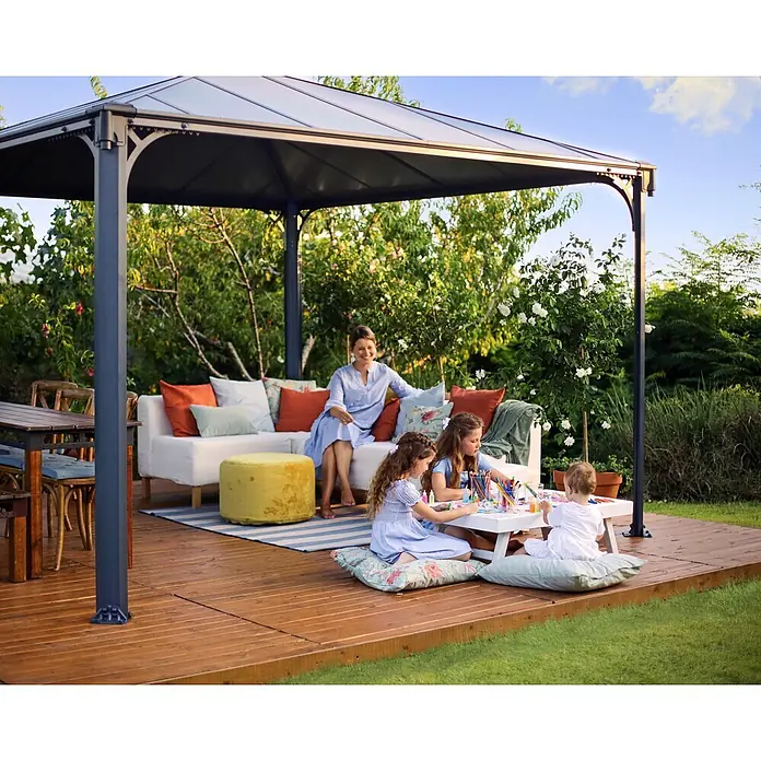 Palram – Canopia Pérgola estándar Palermo - An x Pr: 360 x 360 cm, Material del armazón: Aluminio, Gris - Cenador de metal oscuro con techo sólido, ofrece sombra en una terraza de madera. Una familia se relaja y hace manualidades bajo él en el jardín.