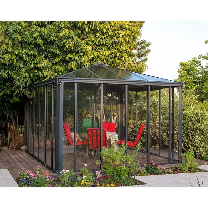 Palram – Canopia Pérgola de jardín cerrado Ledro - An x Pr: 360 x 360 cm, Color del armazón: Antracita, Aluminio - Cenador de cristal con marco gris oscuro, paneles y techo transparentes, sobre tarima de madera en jardín, con sillas rojas y mesa.