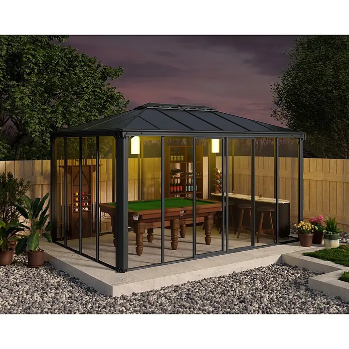 Palram – Canopia Gartenpavillon Ledro 4300 - B x T: 295,5 x 430 cm, Aluminium, Anthrazit - Ein dunkelgerahmter Glaspavillon mit Billardtisch und Bar, von innen beleuchtet, steht im Garten bei Dämmerung.