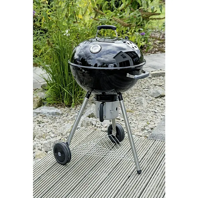 Kingstone Barbacoa redonda con tapa Bullet 57 - Diámetro de la superficie de la parrilla: 57 cm, Negro - Una barbacoa redonda de carbón con termómetro de tapa está en un patio, ideal para parrilladas acogedoras al aire libre.