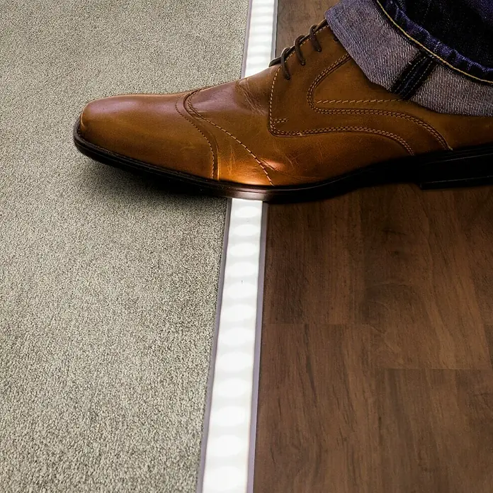 Paulmann Floor Profil Diffusor - 1 m, Aluminium - Das Bild zeigt ein LED-Bodenprofil zwischen Teppich und Holzboden, beleuchtet einen braunen Lederschuh und sorgt für stilvolle Übergänge.