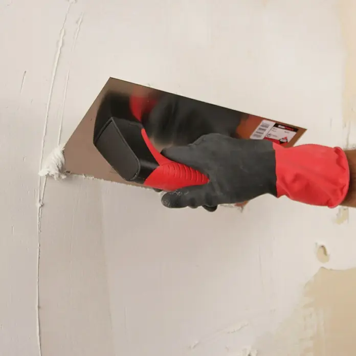 Rubi Llana de alisado Rubiflex - 150 x 300 mm - Un hombre usa una llana para aplanar una pared, asegurando una superficie uniforme. Esta herramienta es esencial para un enlucido perfecto.