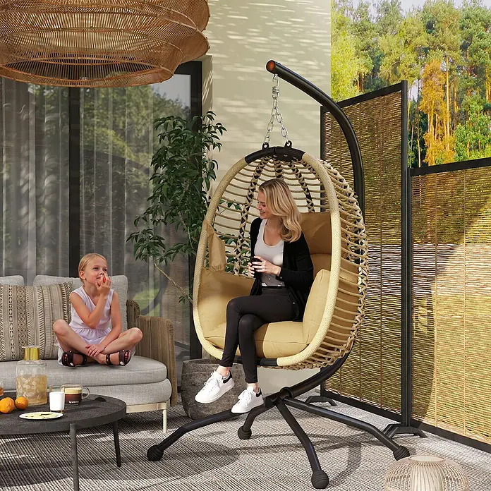 Outsunny Hängesessel - L x B x H: 110 x 100 x 188 cm, Beige - Hängesessel aus Korbgeflecht mit beigefarbenem Kissen und schwarzem Gestell, bequem besetzt von einer Frau auf einer Terrasse.
