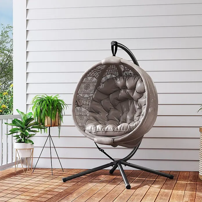Outsunny Hängesessel - L x B x H: 125 x 116 x 172 cm, Sand - Ein grauer eiförmiger Schaukelstuhl mit weichem Kissen und schwarzem Metallgestell steht auf einer Holzterrasse, umgeben von Topfpflanzen.