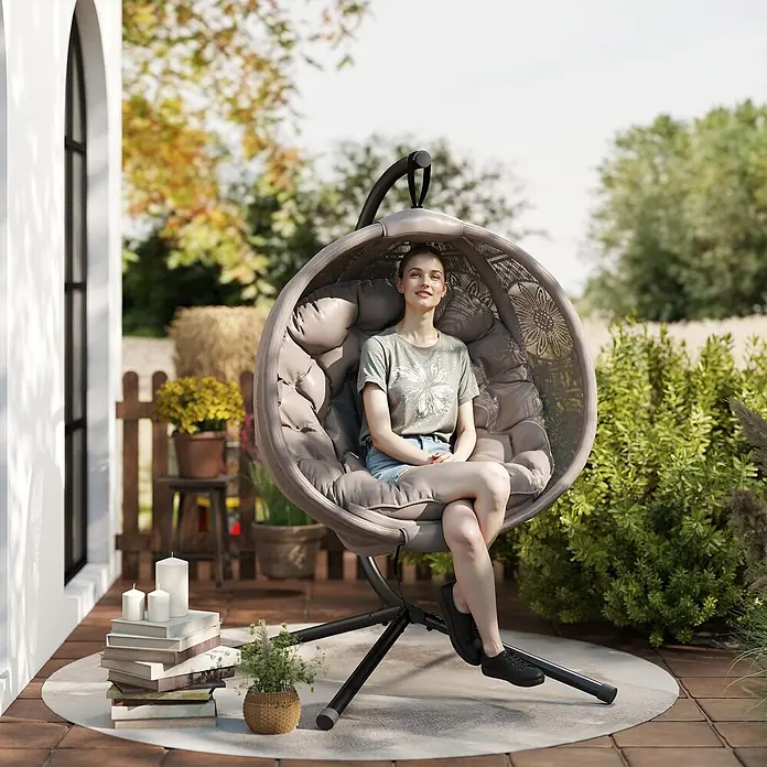 Outsunny Hängesessel - L x B x H: 125 x 116 x 172 cm, Sand - Beiger Hängesessel aus Rattan mit dunklem Metallgestell und weichen Kissen, draußen auf einer Terrasse von einer Frau genutzt.