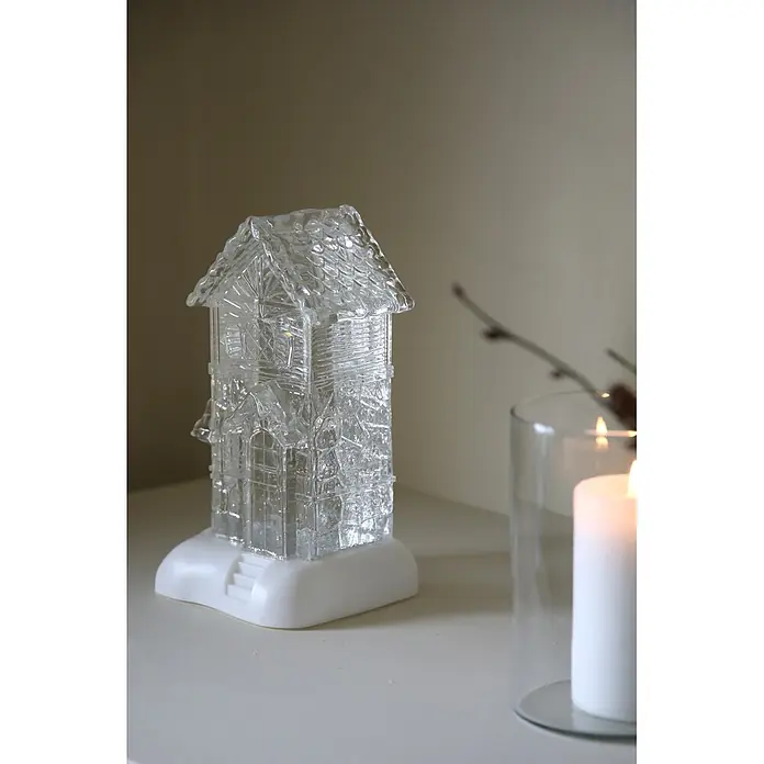 Markslöjd LED-Weihnachtsdekoleuchte Dale - 14,5 x 13 x 26 cm, Weiß/ Transparent, Batteriebetrieben, LED - Klares, hausförmiges Dekorationsstück auf weißem Sockel, neben einer brennenden Kerze.