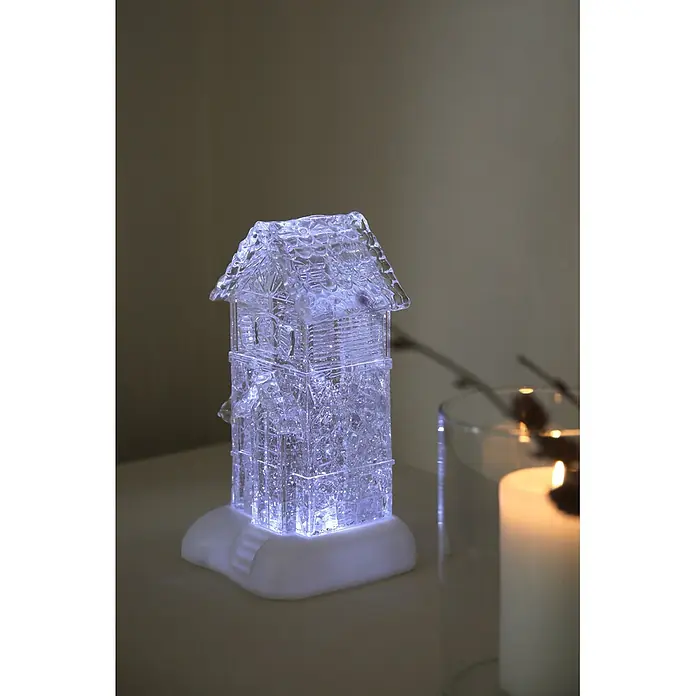 Markslöjd LED-Weihnachtsdekoleuchte Dale - 14,5 x 13 x 26 cm, Weiß/ Transparent, Batteriebetrieben, LED - Ein klares, hausförmiges Acryl-Lichtobjekt, das in kühlem Blau leuchtet, auf einem weißen Sockel.