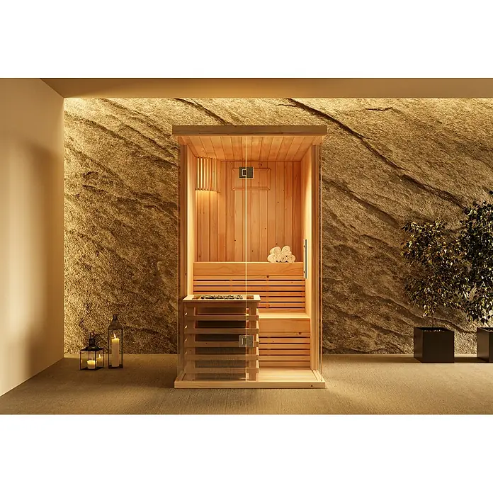 FinnTherm Massivholzsauna Milla -  - Eine moderne Holzsauna-Kabine mit Glasfront, ausgestattet mit internem Ofen und gerollten Handtüchern, in einem Raum mit strukturierter Wand.