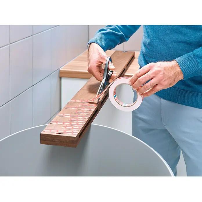 Tesa Powerbond Cinta de doble cara Azulejos/Metal - L x An: 5 m x 1,9 cm, Blanco - Hombre cortando cinta de montaje Tesa con tijeras sobre una tabla de madera sobre un cubo.