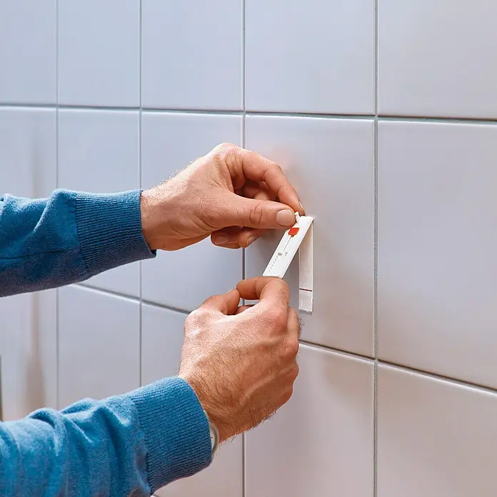 Tesa Clavo adhesivo ajustable - Blanco, Carga soportada: 3 kg, 2 ud. - Una persona que fija un gancho Tesa Powerstrips a una pared de azulejos. El gancho es blanco con un detalle rojo.