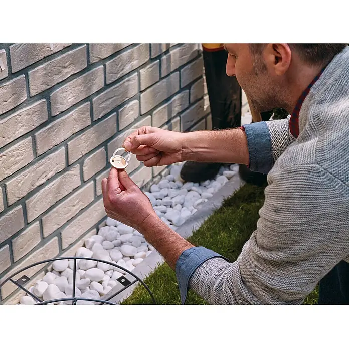 Tesa Tornillo adhesivo - Redonda, Carga soportada: 10 kg, 2 ud. - Un hombre sujeta un sensor de riego a una pared de ladrillos con guijarros blancos y hierba verde al fondo.