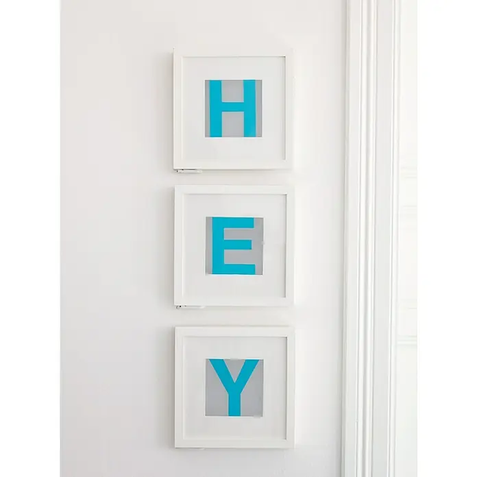 Tesa Clavo adhesivo ajustable - Blanco, Carga soportada: 2 kg, 2 ud. - Una decoración de pared con tres marcos blancos que muestran las letras "H", "E" e "Y" en color turquesa sobre un fondo gris.