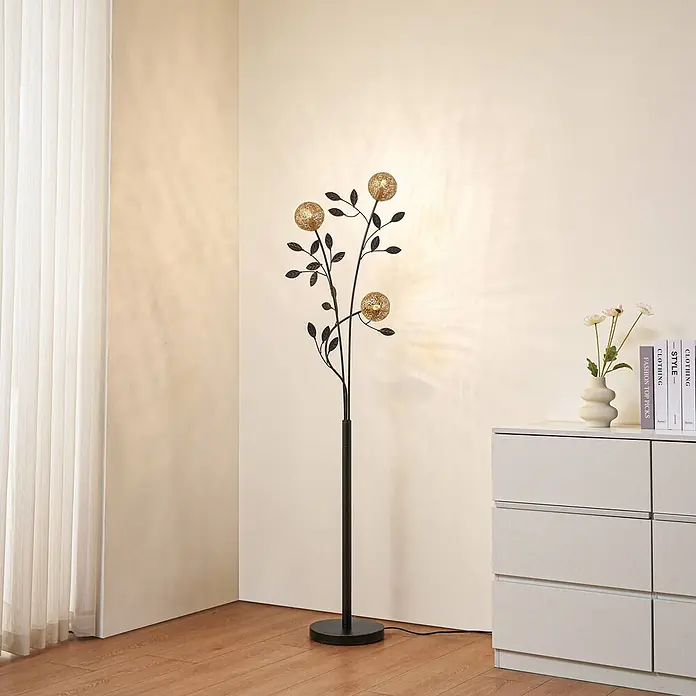 Lucande Stehleuchte Evory - schwarz, messing / gold, G9 - Eine hohe, dunkle Stehlampe aus Metall im Baumdesign mit goldenen Kugelleuchten und dunklen Blättern, die einen modernen Wohnraum beleuchtet.