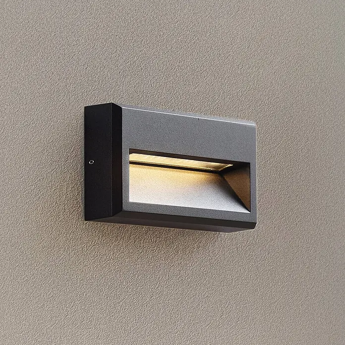 Lucande Außenwandleuchte Weno - 3,5 x 22,7 x 12,7 cm, schwarz, IP65 - Dunkelgraue rechteckige Außenwandleuchte, warm leuchtend, an einer strukturierten Wand montiert.
