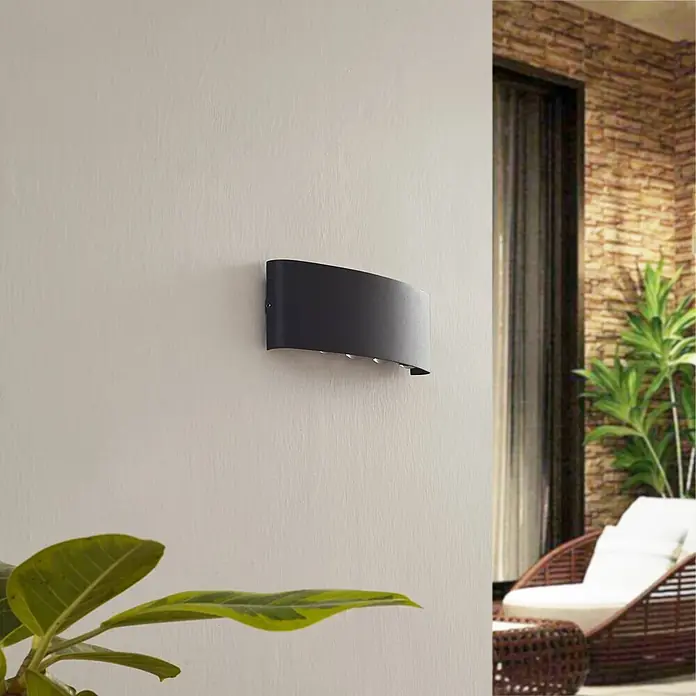 Lindby Außenwandleuchte Ithan - schwarz, IP54 - Schwarze, gebogene Wandleuchte beleuchtet eine Wand in einer Innen-/Außenumgebung.