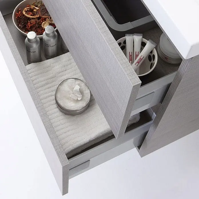 Kiamami Valentina Badmöbel-Set - Grau - Geöffnete hellgraue Badezimmerschubladen mit Holzmaserung, gefüllt mit Handtüchern, Toilettenartikeln und Potpourri.