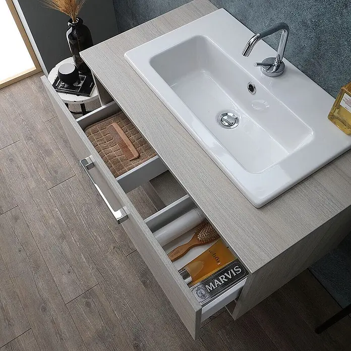Kiamami Valentina Badmöbel-Set - Beige - Badezimmerschrank in heller Holzoptik mit weißem Keramikwaschbecken und zwei offenen Schubladen für Toilettenartikel.