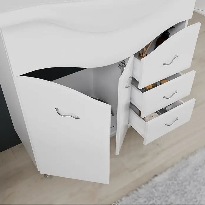 Kiamami Valentina Badmöbel-Set - Weiß - Weißer Badunterschrank mit geschwungener Spüle, offener Tür und drei offenen Schubladen mit Toilettenartikeln, auf hellem Holzboden.