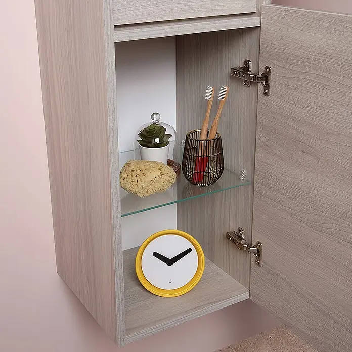 Kiamami Valentina Badmöbel-Set - Helles Holz - Hellholzfarbener Badezimmerschrank, offen, mit Glasablage, Toilettenartikeln, Pflanze und gelber Uhr.