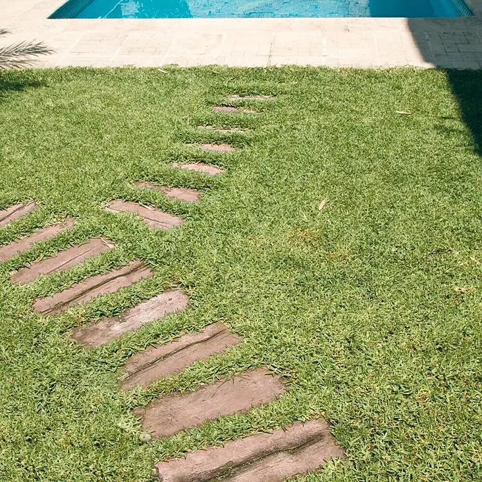Traviesa de madera - 90 x 25 x 5 cm, Hormigón - Un sendero de losas de madera serpentea a través de un césped verde que conduce a una piscina, creando una conexión atractiva y natural.