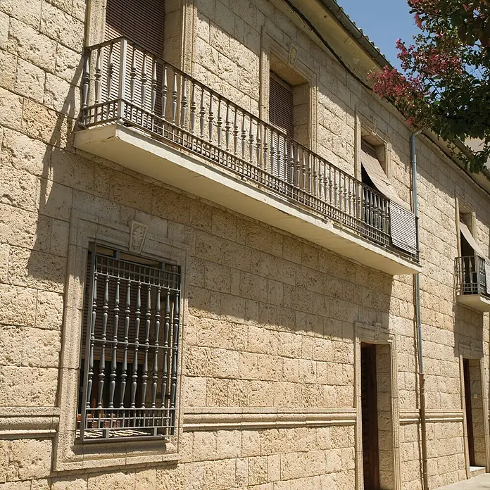 Revestimiento de piedra Coral Piedra - Coral - Vista de una casa color arena con rejas decorativas en las ventanas, barandillas de balcón de hierro forjado y una fachada de piedra rústica.